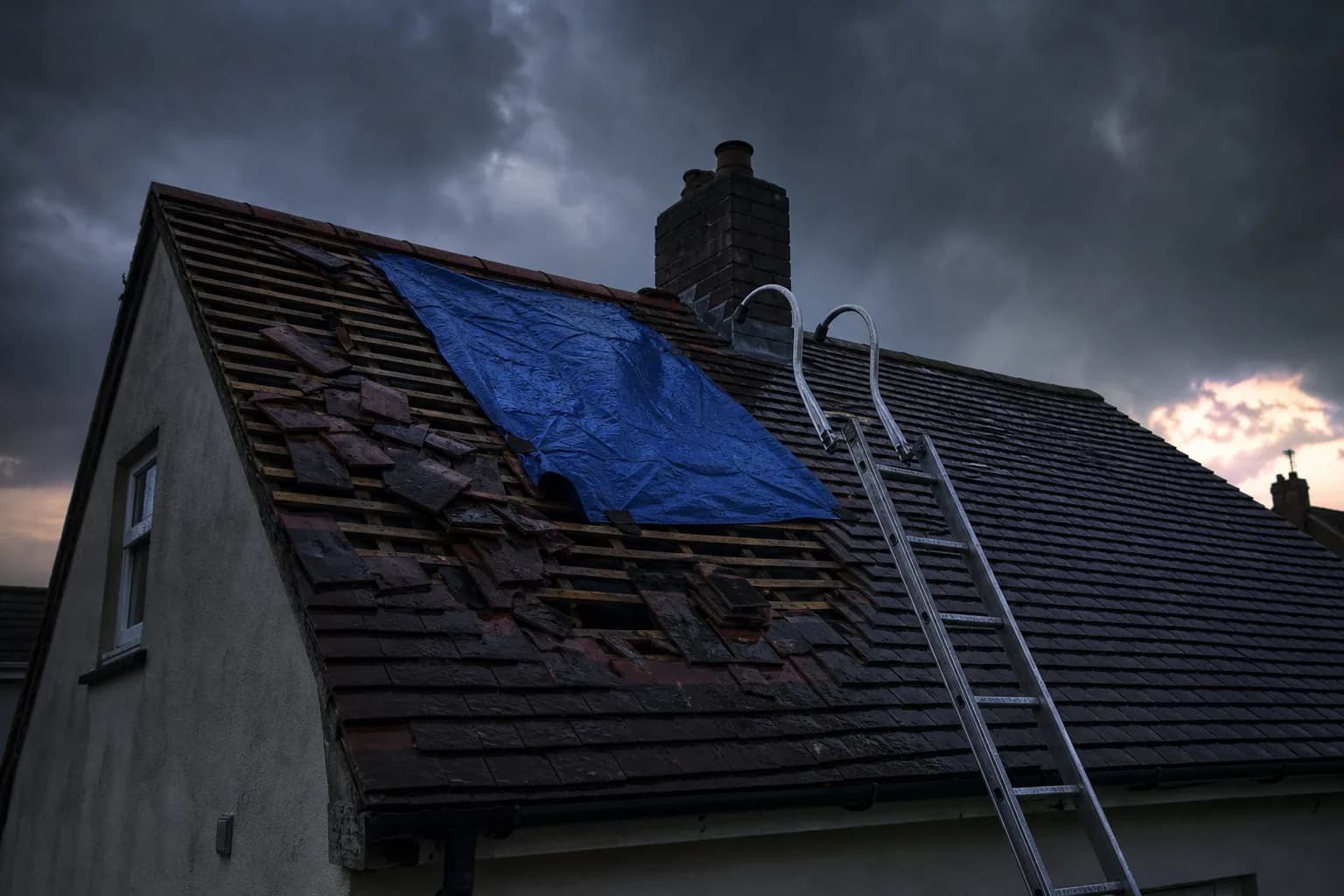Storm-damaged pitched roof with slipped tiles and a tarpaulin under a dark sky