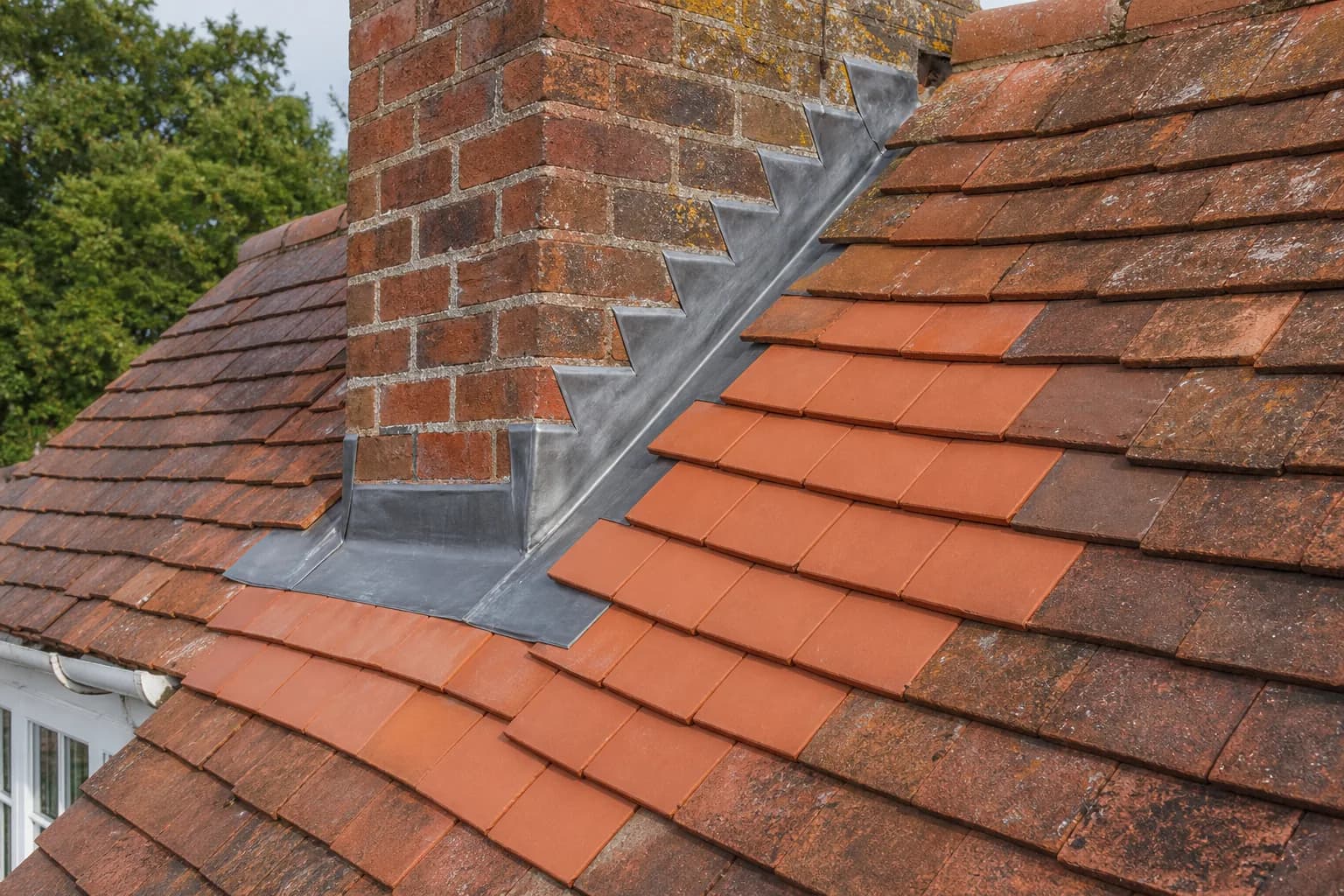 Repaired section of a clay-tiled roof with new tiles and fresh lead flashing around a chimney